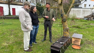 3 Personen stehen im Garten vor der Waschbären-Falle und unterhalten sich: Von links nach rechts Bernd Dauner, Monika Dauner und der Stadtjäger Julian Moosbauer.