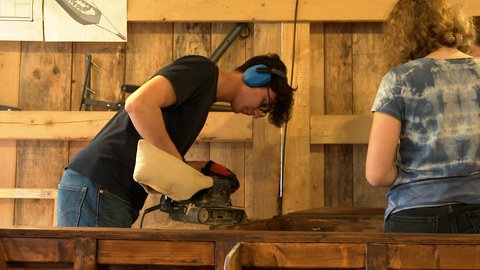 Ein Schüler hat einen blauen Hörschutz auf und schleift mit einer Schleifmaschine ein Stück Holz ab. 