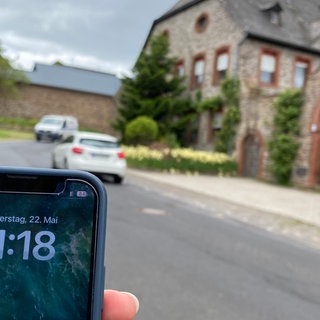 Ein Handy mit schlechtem Empfang, dahinter eine Dorfstraße. (Bürger und Bürgerinnen müssen in Münstermaifeld-Mörz (Kreis Mayen-Koblenz) oft auf der Suche nach Mobilfunkempfang gehen.)