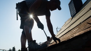 Ein Dachdecker steht bei gleißender Sonne auf dem Dach und arbeitet. Zunehmende Hitze trifft viele Branchen. Die Krankheitstage im Sommer nehmen auch in RLP stark zu.