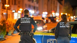 Eine Polizistin und ein Polizist stehen vor ihrem Polizeiwagen mit dem Rücken zur Kamera in einer Wohngegend. 