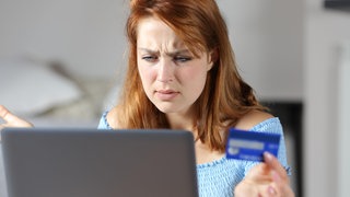 Frau sitzt mit Bankkarte in der Hand irritiert vor dem Laptop
