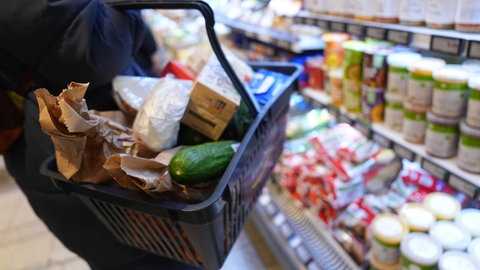 Eine Kundin geht mit einem Einkaufskorb und ihrem Einkauf an den Regalen in einem Supermarkt vorbei.