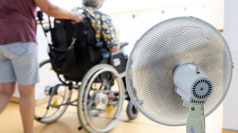Eine Mitarbeiterin eines Seniorenheims schiebt eine Bewohnerin über den Flur, während ein Ventilator einen kühlen Luftzug erzeugt.