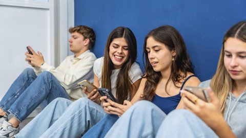 Schülerinnen und Schüler sitzen mit einem Handy vor einer Wand in der Schule.