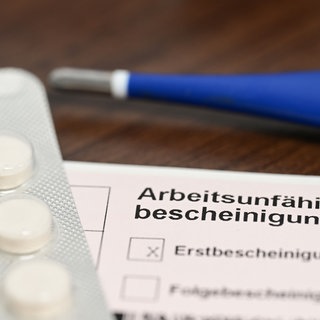 Ein Fieberthermometer, Medikamente und eine ärztliche Arbeitsunfähigkeitsbescheinigung liegen auf einem Nachttisch. Im ersten Halbjahr 2026 haben sich weniger DAK-Versicherte in Rheinland-Pfalz krankschreiben lassen (Symbolbild).