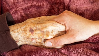 Eine junge Frau hält die Hand einer alten Frau, deren Hand deutliche Altersflecken aufweist.