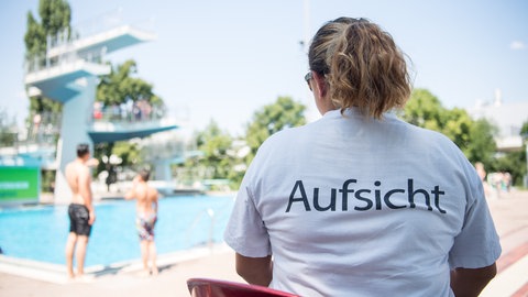 Eine Bademeisterin sitzt am Beckenrand und beobachtet ein Schwimmbecken mit Sprungturm.
