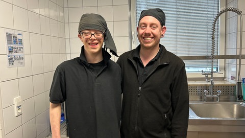 Werkstattbeschäftigte Quentin und Marcel stehen lächelnd nebeneinander von der Kantine im SWR Studio Kaiserslautern.