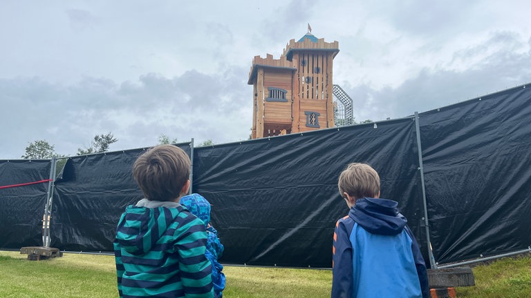 Kinder warten vor einem Bauzaun hinter dem ein Klettergerüst zu sehen ist: Der Unisport der Rheinland-Pfälzischen Technischen Universität Kaiserslautern-Landau (RPTU) hat auf dem Campus einen Abenteuerspielplatz eröffnet. Das Kitz-Land können Kinder mit einer Unisport-Karte besuchen.