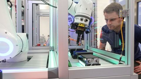 Mehr Start-ups für die Westpfalz, dieses Ziel verfolgt das Netzwerk "Southwest X". In der Smart Factory Kaiserslautern wird Innovationsfreude schon heute gelebt: Der Forschungsverein entwickelt gemeinsam mit Unternehmen Systeme für automatisierte Produktionsprozesse. Eine Produktionsanlage mit Greifarm im Vordergrund, im Hintergrund beobachtet ein Mitarbeiter des Forschungsvereins Smart Factory Kaiserslautern die Abläufe.