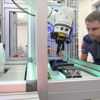 Mehr Start-ups für die Westpfalz, dieses Ziel verfolgt das Netzwerk "Southwest X". In der Smart Factory Kaiserslautern wird Innovationsfreude schon heute gelebt: Der Forschungsverein entwickelt gemeinsam mit Unternehmen Systeme für automatisierte Produktionsprozesse. Eine Produktionsanlage mit Greifarm im Vordergrund, im Hintergrund beobachtet ein Mitarbeiter des Forschungsvereins Smart Factory Kaiserslautern die Abläufe.