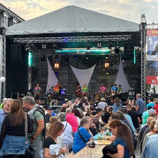 In Zweibrücken wurde am Wochenende das Stadtfest gefeiert. Auf dem Herzogplatz war viel los. Viele Leute sitzen auf Bierbänken vor der großen Bühne, auf der eine Band spielt.