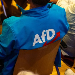 (Symbolbild) Menschen sitzen bei einer Veranstaltung der AfD auf Stühlen, jemand trägt eine blaue Jacke mit einem AfD-Logo.