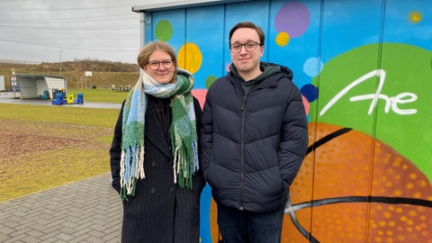 Eine junge Frau und ein junger Mann stehen vor einem bunt bemalten Container auf dem Schulgelände des Are-Gymnasiums in der Grafschaft. 