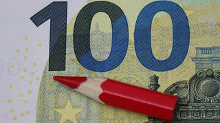 auf einem 100-Euroschein liegt ein roter Stift: Der Rhein-Lahn-Kreis hat für 2025 noch keinen genehmigten Haushalt und muss bei Schulen, Straßen und Vereinen sparen.