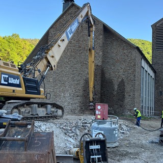 Ein Bagger reißt die St. Andreas Kirche in Ahrbrück ab, ein Bauarbeiter hält einen Wasserschlauch auf die Abrissstelle, um die Staubbildung zu mindern.