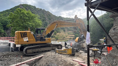 Eine Baumaschine arbeitet im Ahrtal, umgeben von grünen Hügeln, um Gleise zu verlegen.