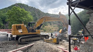 Eine Baumaschine arbeitet im Ahrtal, umgeben von grünen Hügeln, um Gleise zu verlegen.