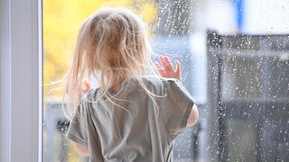Ein Kind steht an einem Fenster mit Regentropfen
