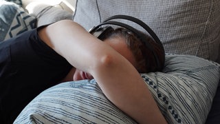 Eine Frau liegt im Bett mit Kopfhörern gegen Geräusche. Menschen, die an den schweren Folgen einer Corona-Infektion leiden, sind geräuschempfindlich und oft isoliert. 