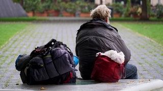 Ein obdachloser Mann sitzt mit dem Rücken zur Kamera auf einer Bank in einem Park. (Symbolbild)