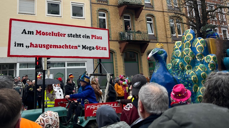 "Am Moselufer steht ein Pfau im hausgemachten Mega-Stau" steht auf einem großen Schild, ein Riesnpfau folgt dahinter.