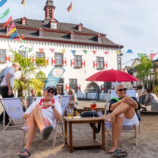 Zwei Personen sitzen entspannt auf einem Liegestuhl auf dem Sandstrand in der Altstadt von Linz am Rhein. 