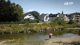Viele Menschen im Ahrtal leiden noch immer unter den Folgen der Flutkatastrophe im Ahrtal. Eine Frau steht an einem heißen Tag mit Schirm an der Ahr.
