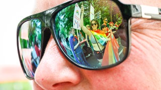 Person mit Sonnenbrille, in der sich Teilnehmer am CSD spiegeln, Symbolbild für queere Menschen zeigen sich seltener öffentlich 