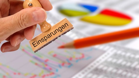 Eine Hand hält einen Stempel mit der aufschrift "Einsparungen": Die Aufsichtsbehörde hat den Haushalt der Stadt Mainz für dieses Jahr genehmigt. 