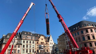 Die Heunensäule am Markt in Mainz schwebt an Kränen über dem Platz. Der Untergrund der Säule muss saniert werden
