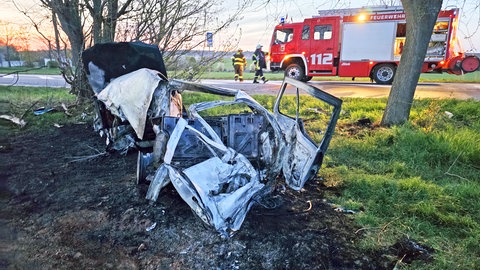 Das ausgebrannte Fahrzeug steht auf einer Wiese im Hintergrund steht ein Löschfahrzeug der Feuerwehr. 