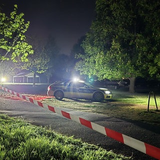 Ein Polizeiauto steht im dunklen Hartenbergpark. Der ist mit einem rot-weißen Flatterband abgesperrt.