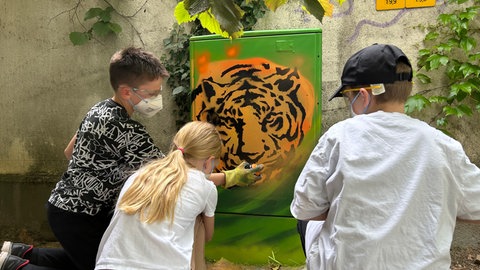 Drei Kinder hocken vor einem Stromkasten. Dieser ist grün und hat einen orange-schwarzen Tigerkopf vorne drauf. 