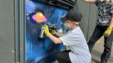 Junge hockt vor Stromkasten und sprüht mit einer Spraydose. 