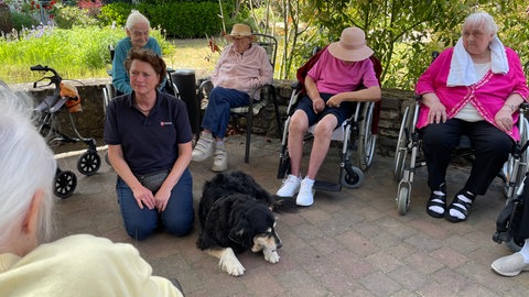 Ehrenamtlerin Birgit Rück kniet mit ihrem Besuchshund Luca zwischen Bewohnerinnen eines Pflegeheims.