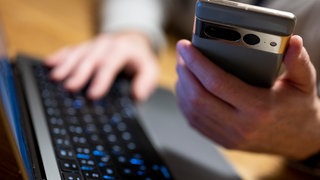 Ein Mann sitzt am Laptop und hält ein Smartphone mit in der Hand. In Mainz haben mehrere Menschen viel Geld an Anlagebetrüger verloren. 