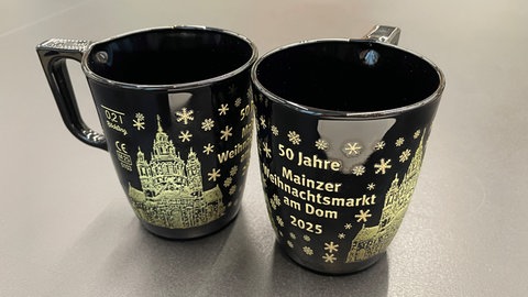 Schwarze Tassen mit goldenem Aufdruck vom Mainzer Dom und der Aufschrift: "50 Jahre Mainzer Weihnachtsmarkt am Dom 2025"