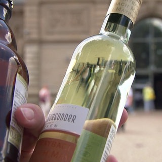 Jemand hält vor dem Mainzer Hauptbahnhof eine Bier- und eine Weinflasche. Es gibt Pläne, Alkohol an Bahnhöfen zu verbieten