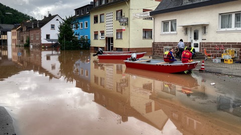 Straße in Kordel (RLP), die nach dem Hochwasser 2021 unter Wasser steht. Am Wasserrand befinden sich drei Boote der Feuerwehr und zwei Einsatzkräfte