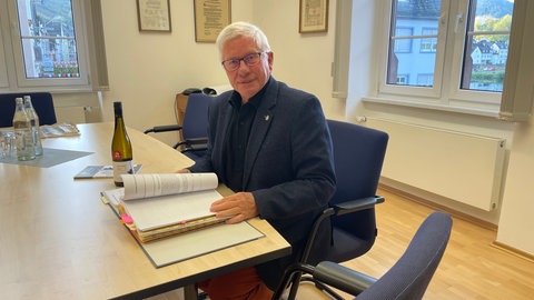 Die Weinlage "Zeller Schwarze Katz" soll wegen des neuen deutschen Weingesetzes umbenannt werden. Die Stadt Zell will sich mit einer Klage gegen die Namensänderung wehren. Der Stadtbürgermeiser von Zell, Hans-Peter Döpgen (FWG), sitzt an seinem Schreibtisch vor einem dicken Ordner voller Schriftverkehr.