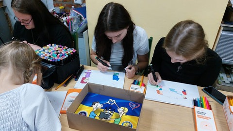 Jugendliche sitzen an einem Tisch einer Schule in der Ukraine und malen Cartoonfiguren mit bunten Farben aus. 
