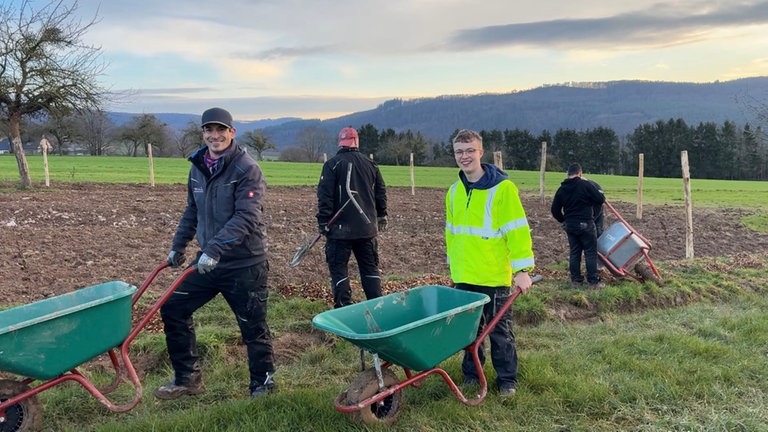 Zwei Menschen schieben leere Schubkarren über eine Wiese vor einem Feld beim Hofgut Serrig.