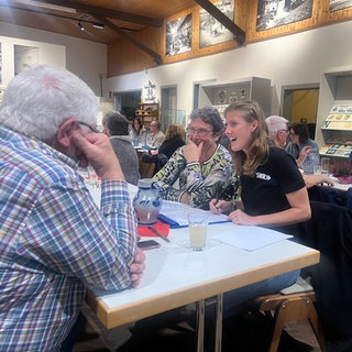 Drei Menschen sitzen an einem Tisch und unterhalten sich, die beiden Frauen sind der Kamera zugewandt, der Mann sitzt mit dem Rücken zur Kamera.
