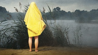 Eine Frau steht barfuß in ein gelbes Handtuch gewickelt am sandigen Ufer eines Flusses vor Schilf und blickt auf den Fluss.