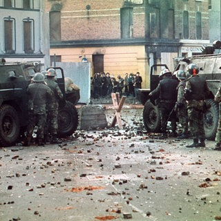 Britische Soldaten mit Panzerwagen stehen auf einer mit Steinen übersäten Straße hinter einer Straßensperre, in einiger Entfernung eine Gruppe von Demonstranten. Am 30. Januar 1972 wurden 13 katholische Demonstranten während einer friedlichen, jedoch verbotenen Kundgebung in der nordirischen Stadt Derry von britischen Fallschirmjägern erschossen.