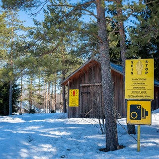 Holzhütte in einem verschneiten Kiefernwald, darin einige gelbe Warnschilder: Grenze zu Russland im finnischen Vaalimaa