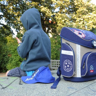 Ein Kind im Kapuzenpulli sitzt mit seinem Schulranzen im Park: Die Gründe für Schulverweigerung sind vielfältig: Häufig haben die Kinder und Jugendlichen Angst vor der Schule, weil sie Mobbing, Stress durch Leistungsdruck oder Probleme mit Lehrkräften erlebt haben. 