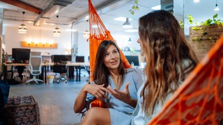 Zwei junge Frauen unterhalten sich in einer Hängematte in einem modernen Büro: Viele der um die Jahrtausendwende Geborenen würden gerne gleich mit einem Teilzeitjob starten. Studien zeigen auch, dass junge Menschen der sogenannten Gen Z Arbeit und Freizeit voneinander trennen und lieber im Büro als im Homeoffice arbeiten möchten. 